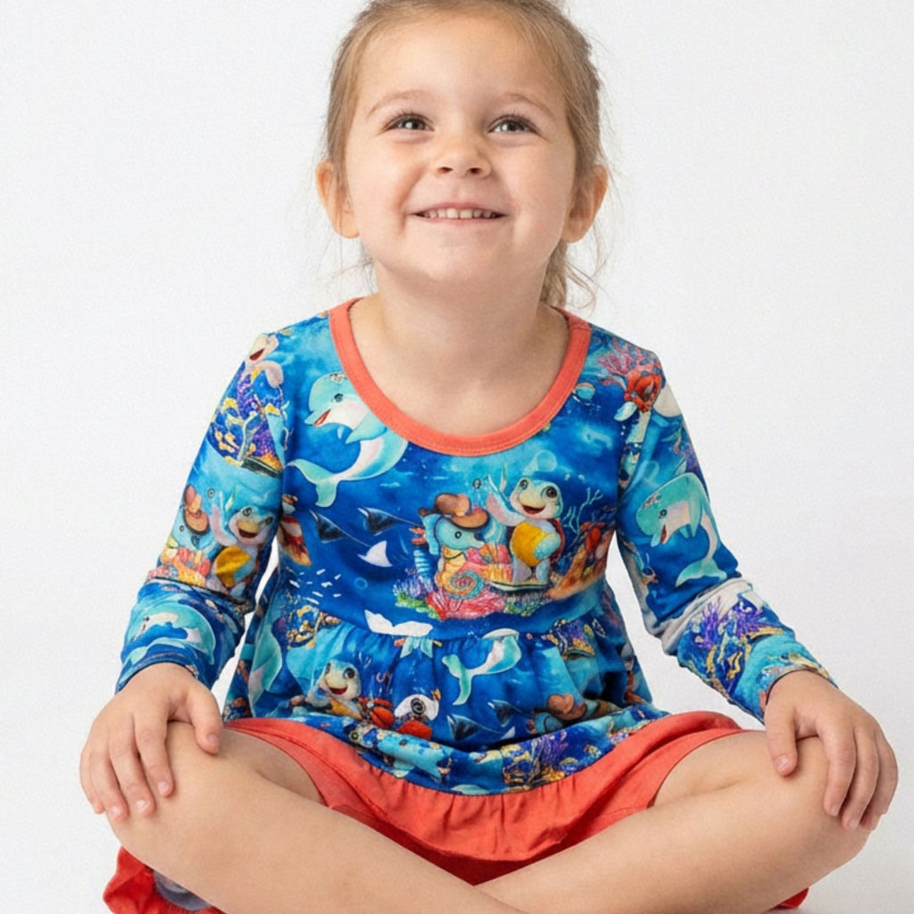 Little girl sitting wearing an ocean-themed bamboo dress with biker shorts by Kai Blue™